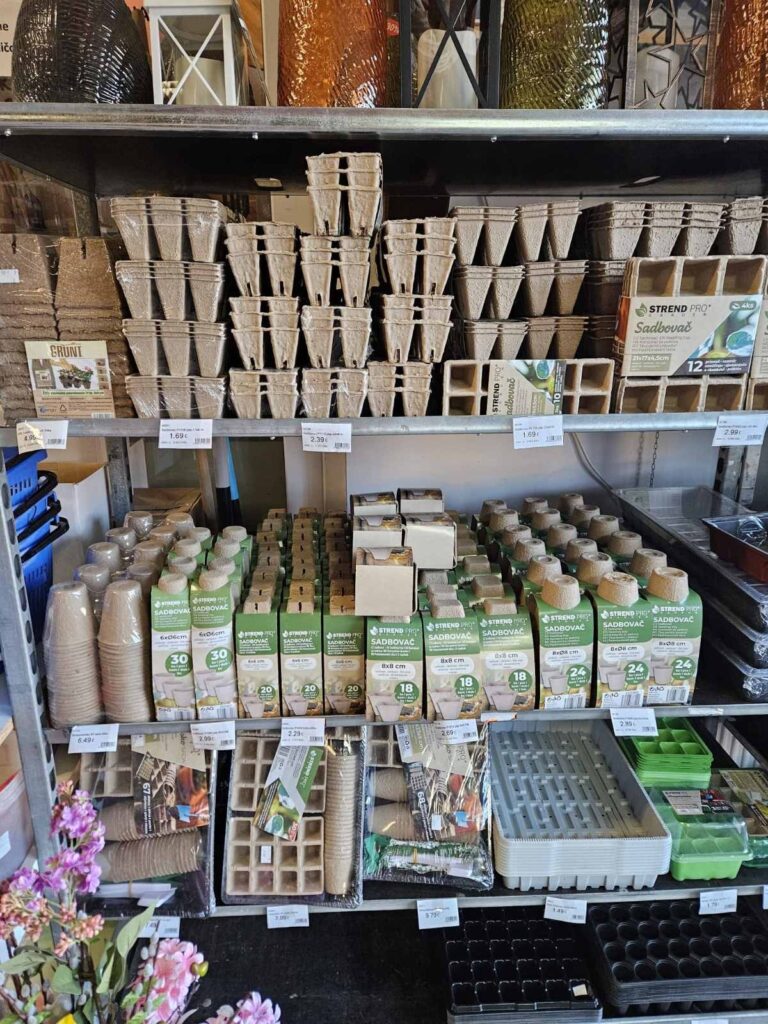 Regál so sadbovými obalmi a rastlinnými nádobami, vrátane papierových kociek a plastových misiek, usporiadaný v záhradníckom obchode.