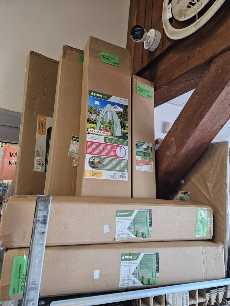 Obrázok zobrazuje niekoľko kartónových obalov s produktmi, pravdepodobne skleníky značky "Strend Pro", umiestnené na regáli v predajni. Na obaloch sú viditeľné cenovky a informácie o rozmeroch produktu.