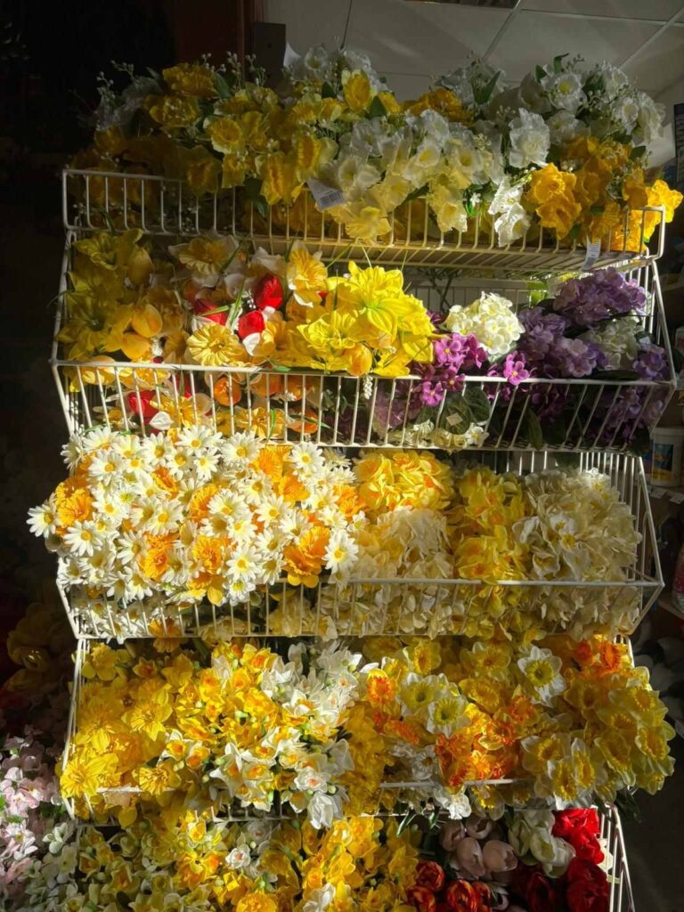 Košíky s umelými kvetmi v rôznych žltých a bielych odtieňoch, usporiadané na policiach.